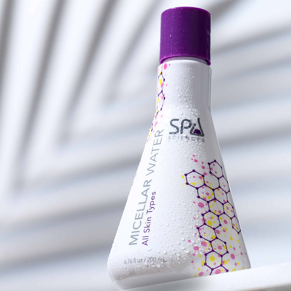 A triangular bottle of Spa Sciences Double Cleanse Duo micellar water with a purple cap, decorated with a yellow and pink hexagonal pattern.
