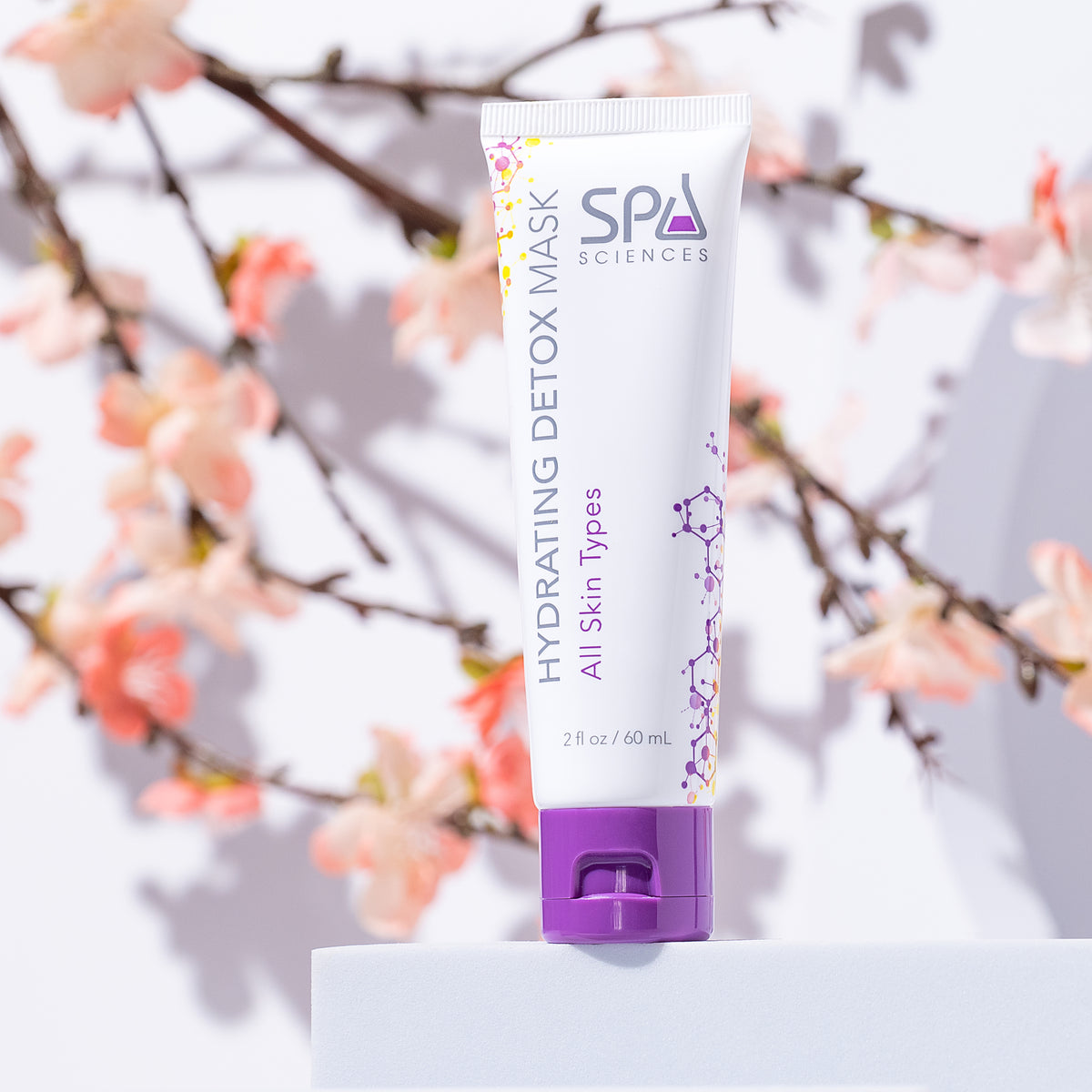 A squeeze tube of Ultimate Self-Care Set hydrating detox mask with a purple cap standing on a white pedestal in front of flowering branches.